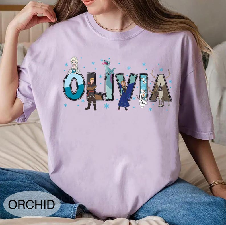 Pu&ograve; includere: Una maglietta lilla chiaro con il nome "OLIVIA" in un carattere decorativo, con personaggi dei cartoni animati e fiocchi di neve. La parola "ORCHID" &egrave; stampata in basso sulla maglietta. La maglietta &egrave; realizzata in un materiale morbido.