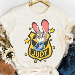 May include: Cream-colored t-shirt featuring a cartoon image of Judy Hopps from Zootopia. Judy, a gray rabbit, is depicted in a police uniform with a badge that reads "JUDY." Yellow stars surround the badge. The shirt is styled with a casual fit.