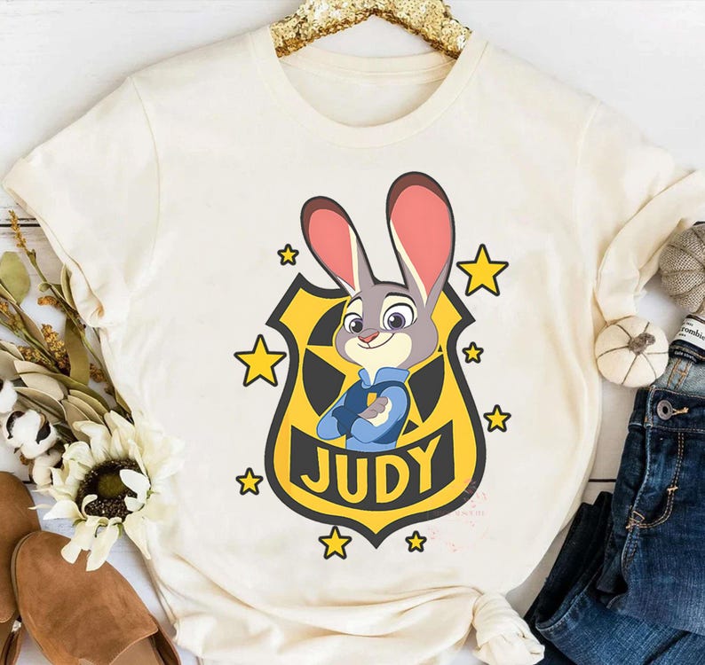 May include: Cream-colored t-shirt featuring a cartoon image of Judy Hopps from Zootopia. Judy, a gray rabbit, is depicted in a police uniform with a badge that reads "JUDY." Yellow stars surround the badge. The shirt is styled with a casual fit.