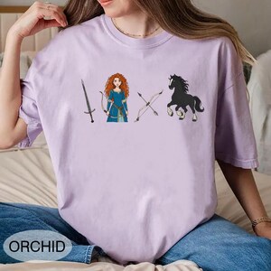 May include: Orchid-colored t-shirt featuring a cartoon illustration of a sword, a character in a blue dress, a bow and arrow, and a black horse. The word "ORCHID" is printed at the bottom. The shirt is made of a soft material.