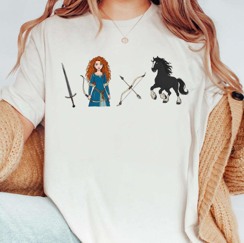May include: Cream-colored t-shirt featuring illustrations of a sword, a woman in a blue dress with a bow and arrow, another bow and arrow, and a black horse. The design has a cartoonish style.