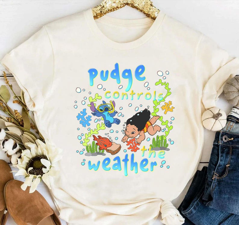 Puede incluir: Camiseta color crema con un gr&aacute;fico colorido. El dise&ntilde;o presenta el texto "Pudge controls the weather", personajes de dibujos animados y elementos submarinos. Es una prenda casual, ideal para el uso diario.