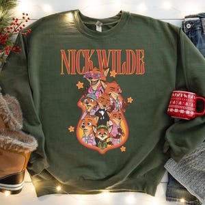 May include: Green sweatshirt featuring a graphic of Nick Wilde from Zootopia, with the text "NICK WILDE" in orange and red. The graphic includes multiple fox characters in various outfits. The sweatshirt is a crew neck style.