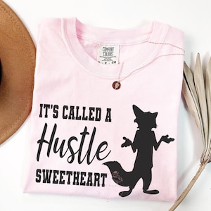 May include: A light pink t-shirt with the text "IT'S CALLED A Hustle SWEETHEART" in black. A black silhouette of a fox is on the right. A gold necklace and a brown hat are visible.