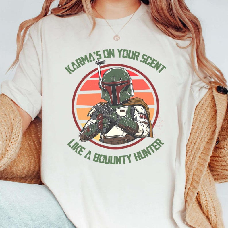 May include: Cream-colored t-shirt featuring a graphic of a bounty hunter with a green and maroon helmet, holding a blaster. The design includes the text "Karma's on Your Scent Like a Bounty Hunter" in green.