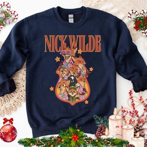 May include: Navy blue sweatshirt featuring a graphic of Nick Wilde and other characters from Zootopia. The text "NICK WILDE" is in orange and yellow. The sweatshirt has a crew neck and long sleeves.