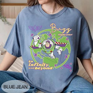 Puede incluir: Una camiseta azul vaquero con un gr&aacute;fico de Buzz Lightyear de Toy Story. El dise&ntilde;o incluye las palabras "Buzz Lightyear" y "to infinity and beyond" en una fuente l&uacute;dica, con estrellas y un gr&aacute;fico de planeta.