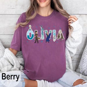 Pu&ograve; includere: T-shirt color bacca con il nome "OLIVIA" in un carattere stravagante, con personaggi dei cartoni animati. Il design include stelle blu e illustrazioni di personaggi. La maglietta ha maniche corte e una vestibilit&agrave; rilassata.