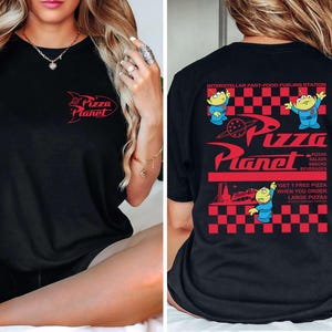 May include: Black t-shirt featuring the Pizza Planet logo in red on the front and back. The back design includes the text "INTERSTELLAR FAST-FOOD FUELING STATION" and cartoon aliens. The shirt also has the text "GET 1 FREE PIZZA WHEN YOU ORDER LARGE PIZZAS."