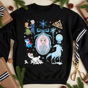 May include: Black sweatshirt with a colourful graphic of characters and symbols from the animated film Frozen. The design includes Elsa, Anna, Olaf, a blue horse, and other elements. The sweatshirt is surrounded by wrapped gifts and pine branches.