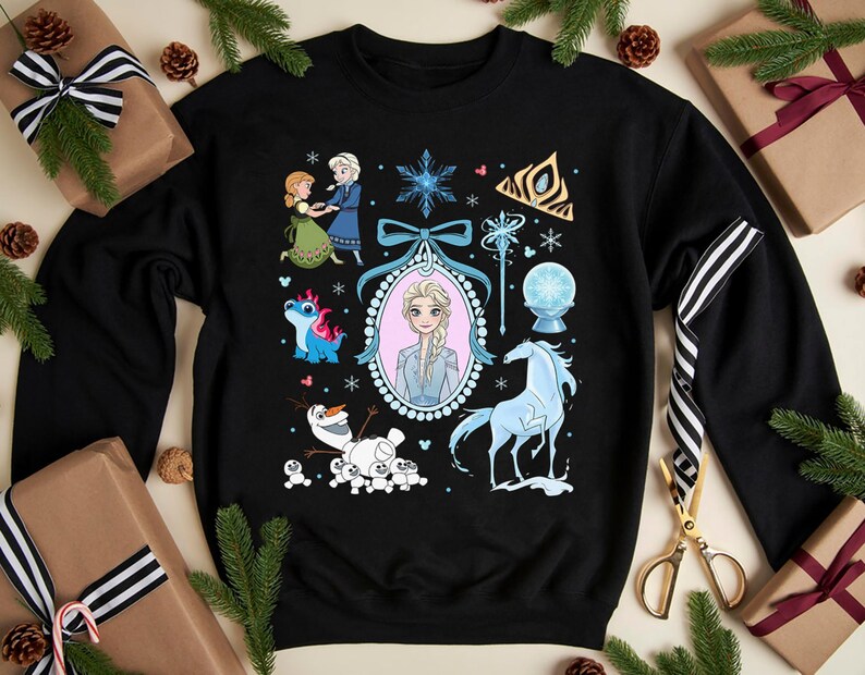 May include: Black sweatshirt with a colourful graphic of characters and symbols from the animated film Frozen. The design includes Elsa, Anna, Olaf, a blue horse, and other elements. The sweatshirt is surrounded by wrapped gifts and pine branches.