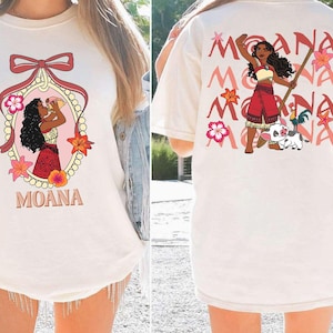 May include: Cream-coloured t-shirt with front and back designs. The front features a cartoon illustration of a woman with the word "MOANA" below. The back displays a similar illustration with the word "MOANA" repeated.