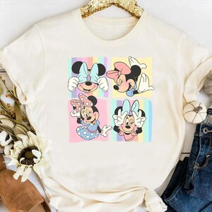 May include: A white t-shirt with a colorful graphic print of four different images of Mickey Mouse. Each image shows Mickey Mouse with a different expression and pose. The background of the graphic is a rainbow.