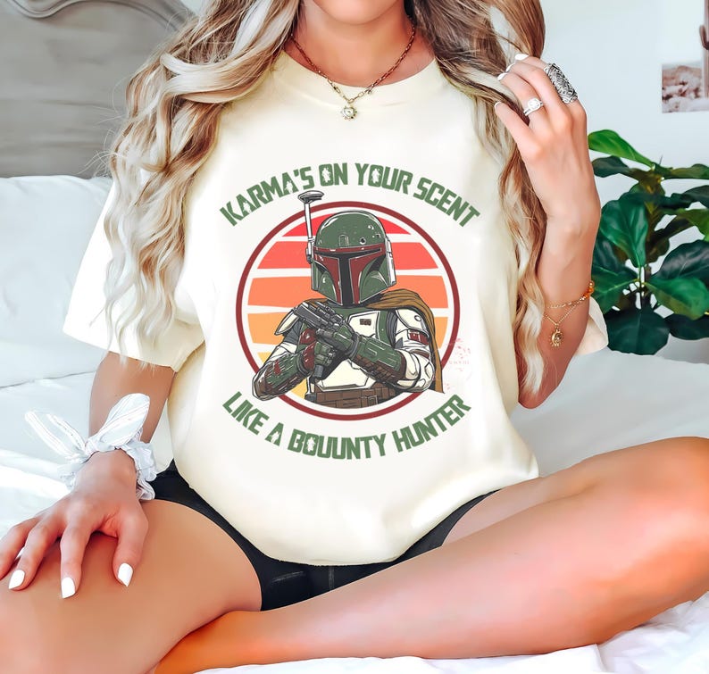 May include: Cream-colored t-shirt featuring a graphic of a bounty hunter in a helmet, set against a sunset-colored circle. The text reads "Karma's on your scent like a bounty hunter."