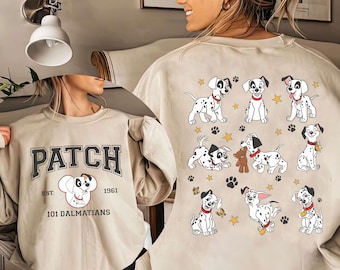 Puede incluir: Sudadera beige con diseño de 101 Dálmatas. La parte delantera muestra "PATCH" en fuente universitaria, un gráfico de dálmata y "EST. 1961". La parte trasera presenta ilustraciones de cachorros dálmatas, huellas de patas y estrellas.