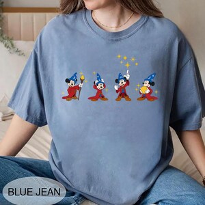 May include: A light blue t-shirt featuring a graphic of Mickey Mouse in a wizard costume. The graphic shows Mickey in four different poses with a red robe, blue hat, and yellow stars. The text "BLUE JEAN" is at the bottom.