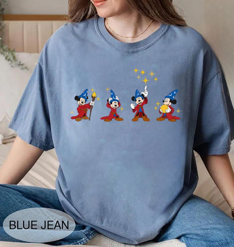 May include: A light blue t-shirt featuring a graphic of Mickey Mouse in a wizard costume. The graphic shows Mickey in four different poses with a red robe, blue hat, and yellow stars. The text "BLUE JEAN" is at the bottom.