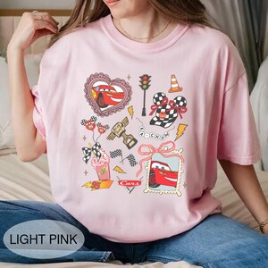 May include: Light pink t-shirt featuring a colorful graphic design with images of a red race car, checkered flags, and a traffic light. The shirt also has the text "Cars" and "95".