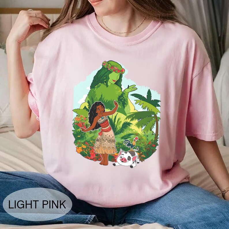 Puede incluir: Camiseta rosa claro con un gr&aacute;fico colorido de una mujer polinesia, una diosa verde, un cerdo y un gallo. El texto "LIGHT PINK" est&aacute; impreso en la parte inferior. La camiseta est&aacute; hecha de un material suave.