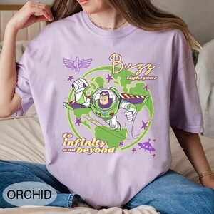 Puede incluir: Una camiseta lila claro con un dise&ntilde;o de Buzz Lightyear. El dise&ntilde;o muestra a Buzz con su traje espacial, las palabras "Buzz Lightyear" y "to infinity and beyond". La palabra "ORCHID" est&aacute; impresa en la parte inferior.
