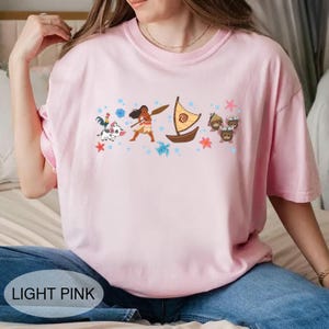 May include: Pale pink t-shirt with a graphic of a Polynesian woman, a boat, and cartoon characters. The shirt has the text "LIGHT PINK" at the bottom. The shirt is made of a soft material.