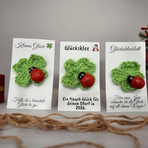 Könnte beinhalten: Drei weiße Karten mit grünen gehäkelten Kleeblättern und roten Marienkäfer-Akzenten. Jede Karte hat deutschen Text. Die Karten sind mit einem braunen Faden verbunden. Die Karten sind für Glück.