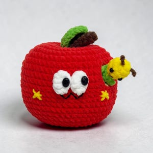 May include: A handmade red crochet apple with white eyes, a smiling mouth, and yellow star accents. A yellow caterpillar with green accents peeks out from a hole in the apple. The apple has a green and brown stem.