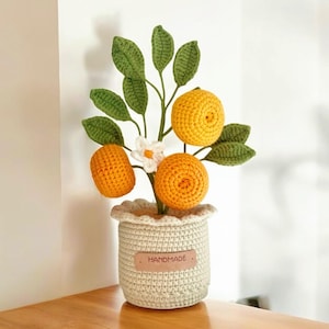 May include: A handmade crochet orange tree in a cream-coloured pot. The tree features bright orange crochet oranges, green leaves, and a white flower. The pot has a "HANDMADE" tag.