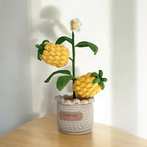 May include: A handmade crochet plant featuring yellow pineapple-shaped fruit and green leaves. The plant is in a grey crochet pot with a "HANDMADE" tag. A white and yellow flower sits atop the plant.
