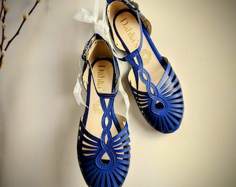 Puede incluir: Un par de sandalias de cuero azul con un diseño tejido y correas de tobillo. Las sandalias tienen el nombre de la marca "Dahlia's" escrito en las correas.