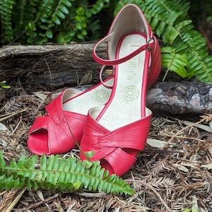 Op de afbeelding: Een paar rode leren peeptoe sandalen met enkelbandjes. De sandalen liggen op een bed van bruine dennennaalden.