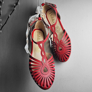 以下が含まれることがあります： 足首に白いリボンが結ばれた赤い革のサンダルです。サンダルはカットアウトデザインになっており、ストラップには「Dahlia」というブランド名が印刷されています。