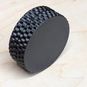 May include: A black, cylindrical object with a textured, honeycomb-like pattern on the side and a smooth, flat surface on the top and bottom. The object is made of a matte material.