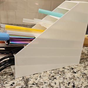 Puede incluir: Un organizador blanco en ángulo lleno de varias pajitas reutilizables. Las pajitas son de diferentes colores, incluyendo negro, amarillo, azul e iridiscente. El organizador está hecho de un material translúcido y se encuentra sobre una encimera moteada.