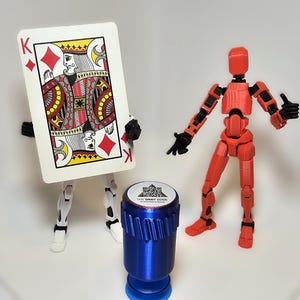 May include: An image featuring a King of Diamonds playing card held by a white and black articulated figure, a red articulated figure, and a blue bottle stopper with a white label.