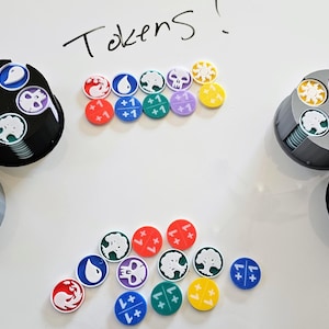 May include: A collection of colorful game tokens and two cylindrical token holders on a white surface. The tokens feature various symbols and colors, including red, blue, green, and yellow. The word "Tokens" is handwritten at the top.