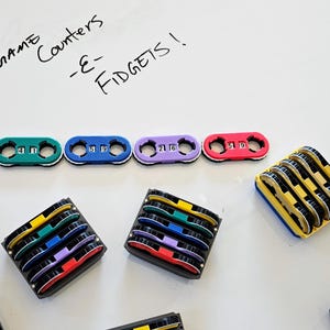 Könnte beinhalten: Bunte Spielzähler und Fidgets. Die Zähler sind gelb, grün, blau, lila und rot, jeweils mit einer Zahlenanzeige. Mehrere Stapel Fidgets in verschiedenen Farben sind ebenfalls vorhanden.