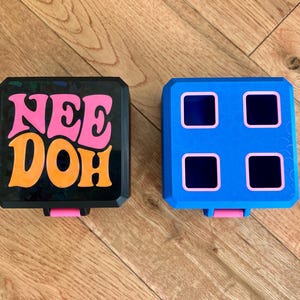 May include: Two square containers with rounded corners. The left container is black with the text NEE DOH in pink and orange. The right container is blue with four square cutouts outlined in pink. Both containers have a small pink tab at the bottom.