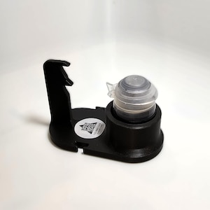 Pode incluir: Um suporte preto para frascos de tinta com um frasco de tinta com tampa transparente. O suporte tem uma pega embutida e uma base circular para segurar o frasco. Um autocolante com texto está na base.