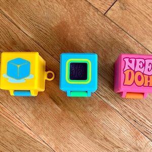 Könnte beinhalten: Drei quadratische Nee Doh Fidget-Spielzeuge in Gelb, Blau und Pink. Das gelbe Spielzeug zeigt eine blaue Eiswürfelgrafik, das blaue Spielzeug hat einen grünen quadratischen Rand und das pinke Spielzeug zeigt den Text Nee Doh in orangefarbenen Buchstaben.