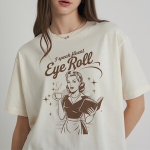 May include: A white t-shirt with a brown graphic of a woman holding a cup of coffee and a book. The text on the shirt reads "I speak fluent Eye Roll".