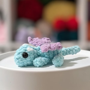 Puede incluir: Un pequeño dragón tejido a ganchillo, en tonos azul claro y lavanda. El juguete hecho a mano presenta un diseño detallado con ojos de seguridad negros y una cola enroscada. El dragón se exhibe sobre una superficie blanca y redonda.