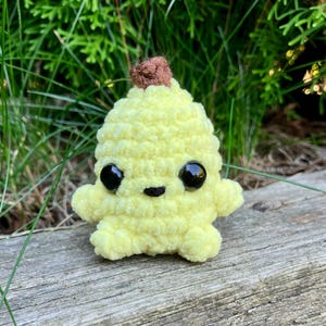 Peut inclure: Une créature jaune en crochet, avec des yeux noirs et un sommet marron. Le jouet en peluche fait main est posé sur une surface en bois vieilli, avec un fond de verdure. La créature a un visage souriant et de petits bras.