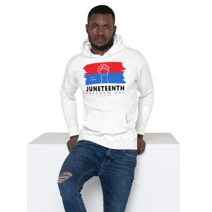 Puede incluir: Sudadera con capucha blanca con un gráfico de Juneteenth. El diseño presenta una pincelada roja y azul con un puño en alto, la palabra "Juneteenth" y "Freedom Day" en texto negro. La sudadera tiene una capucha con cordón y un bolsillo delantero.