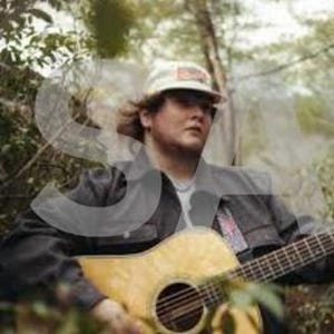 Pode incluir: Uma pessoa usando um boné branco e uma jaqueta cinza está segurando um violão acústico marrom claro. O violão tem uma boca redonda e uma escala escura. O fundo é uma cena externa desfocada com árvores e folhagem.