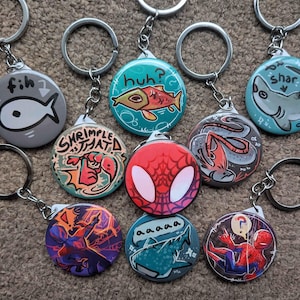May include: A collection of round keychains with various cartoon designs. The keychains feature images of fish, sharks, a spider-man design, and other illustrations. Each keychain has a metal ring and chain for attaching to keys or bags.