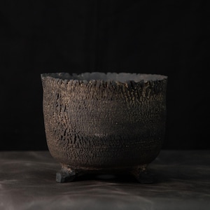 May include: A dark, textured ceramic bowl with a rough, bark-like exterior finish and a deep, rounded basin. The vessel is supported by three small, sturdy feet at the base, featuring a matte, charcoal-colored surface with subtle brown undertones.