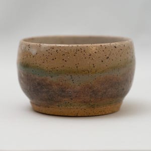 May include: A small, round ceramic bowl with a speckled, earthy design. The bowl features bands of brown, green, and dark brown hues. The interior is a lighter tan color. The bowl has a smooth, rounded shape.