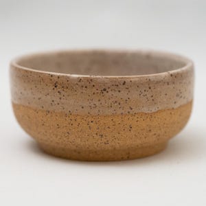 May include: A small, handcrafted ceramic bowl with a speckled, two-tone design. The upper half is a light beige with dark speckles, while the lower half is a warm, earthy brown. The bowl has a rounded shape and a smooth, glazed finish.
