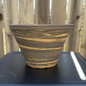 May include: A small, black and brown ceramic pot with a flared rim and a swirling pattern.
